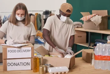 Zwei maskierte Freiwillige packen 'FOOD DELIVERY'- und 'DONATION'-Kartons mit Hilfsgütern.