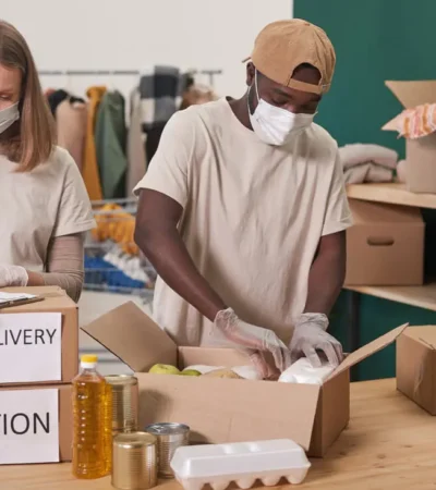 Zwei maskierte Freiwillige packen 'FOOD DELIVERY'- und 'DONATION'-Kartons mit Hilfsgütern.